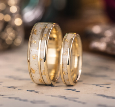A custom gold ring set with beach sand inlay and scattered gold leaf flakes, displayed on a piece of paper with handwritten text and a soft-focus background.