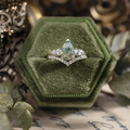 The Liora white gold ring displayed in a green velvet ring box, featuring a pear-shaped moss agate center stone, pear-shaped diamond side stones, and smaller diamond accents underneath.