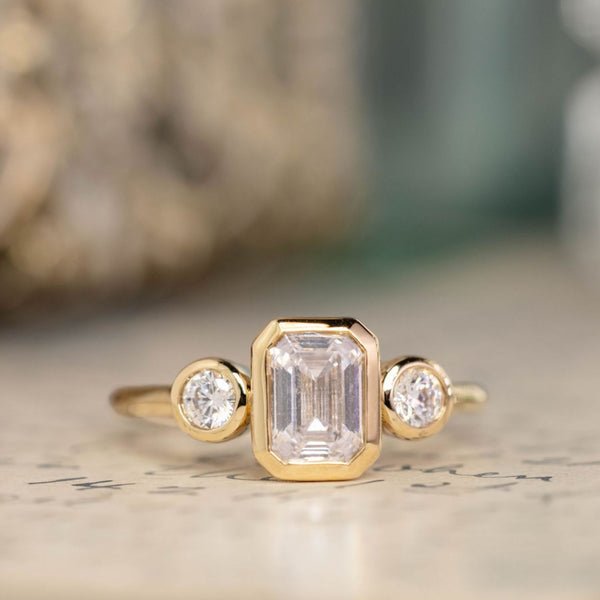 the-marcelline-three-stone-emerald-cut-diamond-engagement-ring-round-diamond-accents-bezel-setting