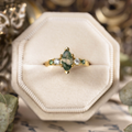 The Titania yellow gold ring displayed in a cream velvet ring box, featuring a marquise moss agate center stone with clear and green accent stones along the band.