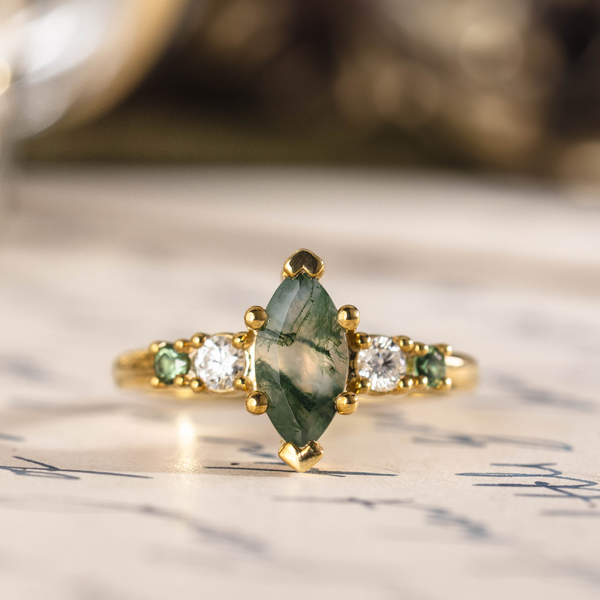 Front view of The Titania yellow gold ring featuring a marquise moss agate center stone with alternating clear and green accent stones on each side.